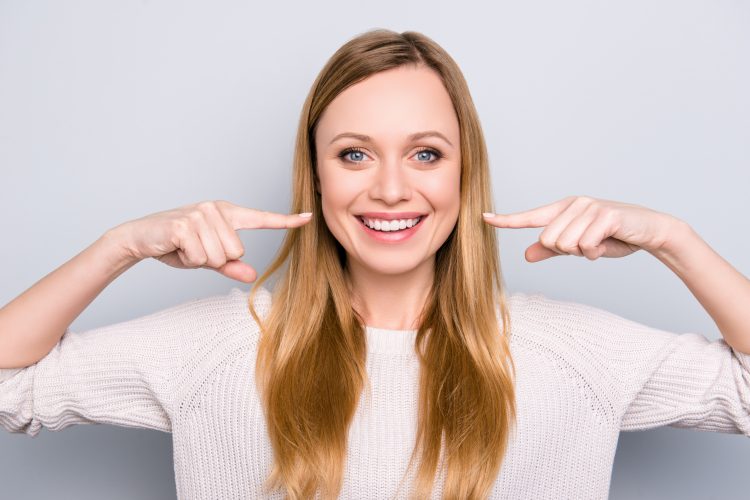 Portrait of joyful satisfied girl gesturing her beaming white healthy teeth with two forefingers looking at camera isolated on grey background. Orthodontic concept